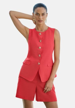 Frau mit dunklen Haaren in einer ärmellosen roten Weste mit Knöpfen und passenden Shorts, die goldene Ohrringe trägt und mit einem Arm hinter dem Kopf posiert.