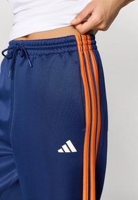 Marineblå joggebukse med tre vertikale oransje striper, elastisk midjebånd med snor, og en hvit logo på venstre lår.