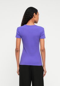 Donna con lunghi capelli ricci, che indossa una maglietta aderente viola a maniche corte e pantaloni neri, voltata di spalle su uno sfondo bianco uniforme.