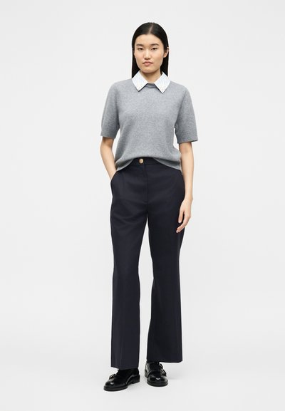 Femme debout devant un fond blanc, portant un pull à manches courtes gris par-dessus une chemise à col, un pantalon noir sur mesure et des chaussures noires.