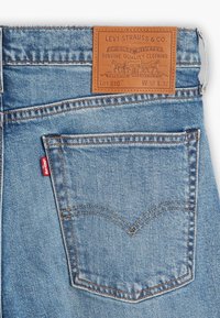 Levi's denimjeans i ljusblått med en brun läderlapp. Funktioner inkluderar stickningar, en röd etikett på fickan och en klassisk design på bakfickan.