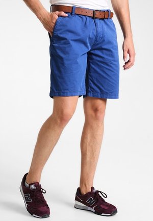 Mann trägt blaue knielange Shorts mit braunem Gürtel und burgunderfarbenen New Balance Sneakers, steht mit einer Hand in der Tasche.