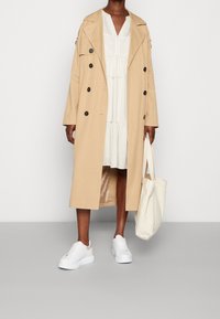 Manteau trench beige à double boutonnage, longueur genou, associé à une robe blanche à rayures verticales et des baskets blanches. Un sac fourre-tout crème est porté.