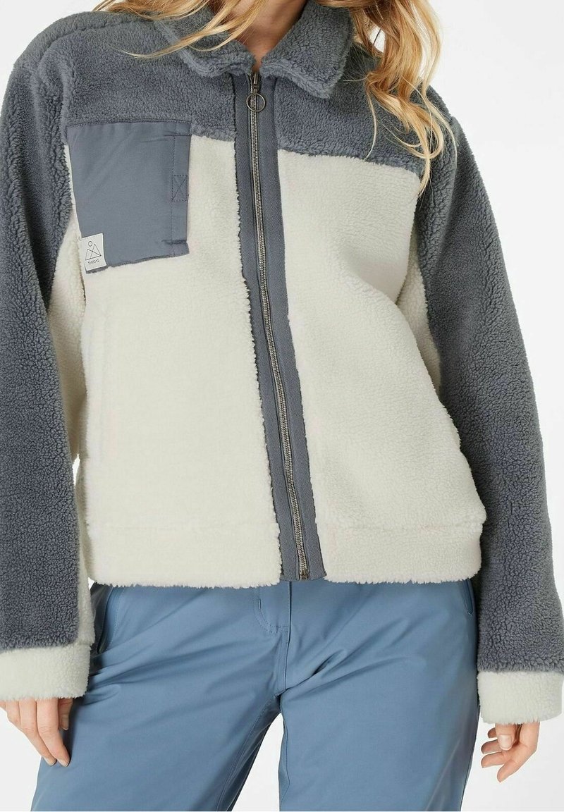 Vrouw in een fleecejack met grijze mouwen en kraag, witte torso, grijze borstzak en blauwe broek, staand met ontspannende handen langs de zij.