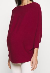 Femme portant un pull ample à manches longues côtelé de couleur rouge foncé et un pantalon clair, avec une boucle d'oreille pendante visible à une oreille.