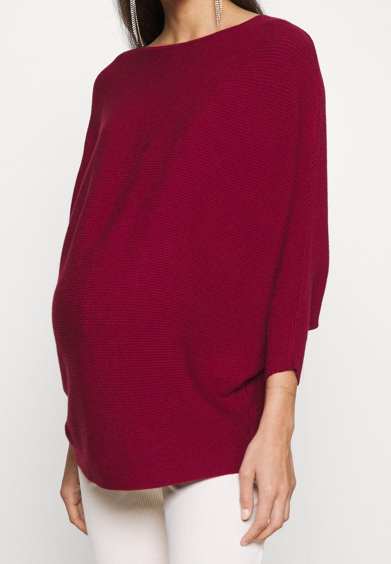 Femme portant un pull ample à manches longues côtelé de couleur rouge foncé et un pantalon clair, avec une boucle d'oreille pendante visible à une oreille.