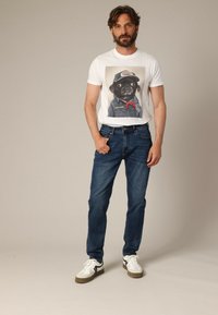 Homme portant un t-shirt blanc avec un graphique de chien en veste en jean et casquette, un jean bleu et des baskets blanches, se tenant contre un fond neutre.
