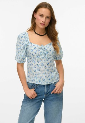 Blusa floreale azzurro chiaro con maniche a sbuffo, scollatura quadrata e dettaglio con nodo frontale. Abbinata a jeans azzurri chiari. Tessuto dalla superficie liscia.