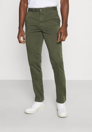 Homme portant un pantalon ajusté vert olive, des baskets blanches et un t-shirt blanc rentré, debout devant un arrière-plan uni.