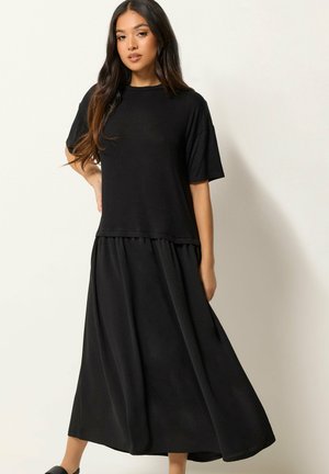 Femme aux longs cheveux foncés portant un t-shirt noir ample et une jupe maxi noire fluide, debout avec une main sur la hanche.
