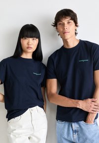 Navy blue cotton t-shirts with small turquoise "tommy jeans" logo. Two models, one wearing white jeans, the other in denim shorts.