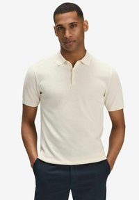 Kortärmad polo skjorta i ljusbeige, med en strukturerad tygkvalitet, treknappslå och klassisk krage. Bärs med mörka byxor.