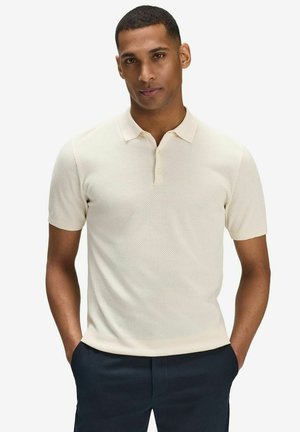 Kortærmet polo shirt i lys beige med struktureret stof, tre-knaps knaplukning og klassisk krave. Båret med mørke bukser.