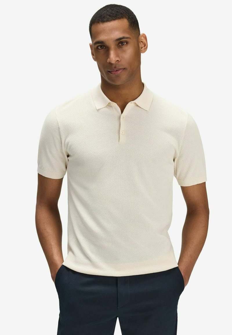 Kortärmad polo skjorta i ljusbeige, med en strukturerad tygkvalitet, treknappslå och klassisk krage. Bärs med mörka byxor.