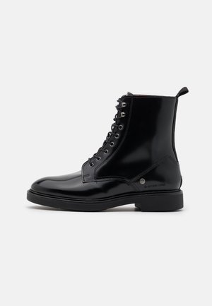 Bottine noire brillante à lacets avec finition en cuir lisse, bout rond, languette à l'arrière et talon bas sur fond blanc.