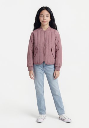 Pige med langt mørkt hår iført en lyngfarvet quiltet jakke, lyseblå jeans og hvide sneakers, stående mod en ensfarvet hvid baggrund.