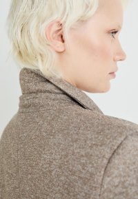 Hnedý štruktúrovaný kabát s vysokým golierom a upraveným strihom, vyrobený z mäkkej látky s jemným vzorom po celom povrchu.