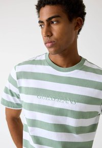Gestreiftes T-Shirt in Grün und Weiß, mit besticktem "GUESS JEANS"-Logo, Rundhalsausschnitt und kurzen Ärmeln. Aus weichem Baumwollmaterial.