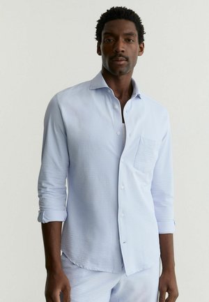 Mand med kort krøllet hår iført en lyseblå tekstureret langærmet skjorte med optrukne ærmer og matchende bukser foran en ensfarvet baggrund.