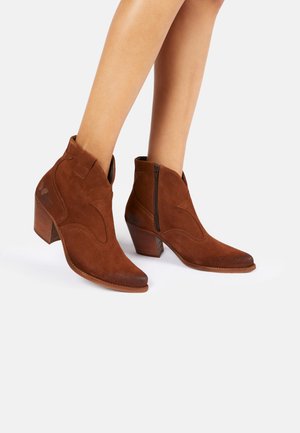 Stivaletti in suede marrone, alla caviglia, con punta affusolata e tacco leggero, caratterizzati da una cucitura decorativa e una cerniera laterale per la chiusura.