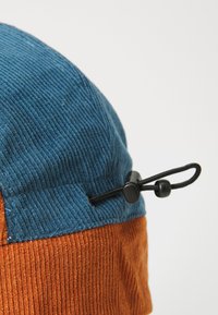 Czapka z sztruksu w niebiesko-pomarańczowym kolorze, z teksturowanym materiałem, kontrastującymi kolorami oraz regulowanym czarnym sznurkiem z zapięciem na plecach.