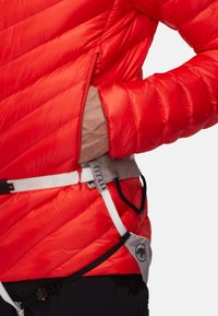 Giacca isolata rossa con design trapuntato, dotata di un colletto alto e polsini elasticizzati, indossata sopra un'imbracatura in vita con cinghie regolabili.