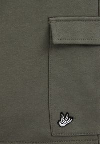 Prim-plan al buzunarului din material verde măsliniu, cu un mic emblemă brodată alb-negru a unei păsări în colțul din dreapta jos.