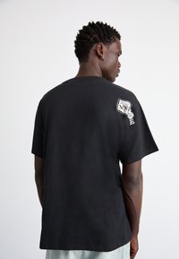 Camiseta de algodón negra de manga corta, que presenta un diseño gráfico en la parte posterior y costura texturizada a lo largo de la costura del hombro.
