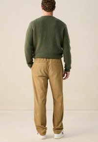 Homme portant un pull vert, un pantalon en velours côtelé beige et des baskets blanches se tenant dos à un fond uni.
