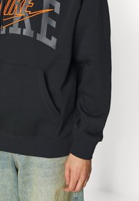Svart hoodie i mjuk tyg, med en stor grå "NIKE"-logotyp och en orange swoosh, samt en framficka och ribbade mudd.