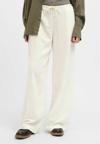 Un pantalon large blanc avec une taille à cordon, confectionné dans un tissu doux. Porté avec un haut gris et une chemise boutonnée vert olive.
