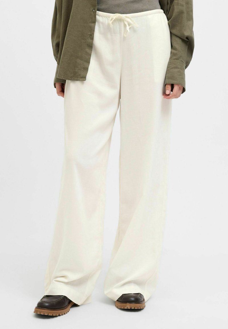 Un pantalon large blanc avec une taille à cordon, confectionné dans un tissu doux. Porté avec un haut gris et une chemise boutonnée vert olive.