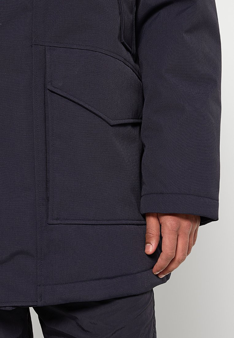 Donkerblauwe thermojack met een gestructureerde stof, een grote zijzak en een manchet aan de mouwen. Het ontwerp heeft versterkte naden.