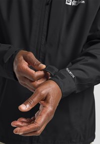 Schwarze wasserdichte Jacke mit strukturiertem Finish, ausgestattet mit verstellbaren Bündchen und einem Markenlogo auf der Brust. Hände, die das Bündchen anpassen.