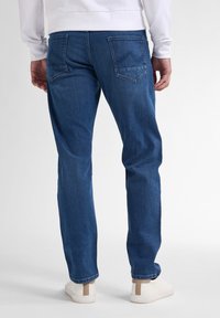 Jeans blu scuro con vestibilità regolare, cuciture visibili, due tasche posteriori e gamba leggermente affusolata, abbinati a scarpe da tennis bianche.