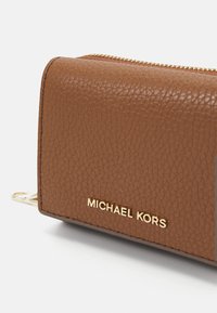 Smeđi teksturirani kožni novčanik s zlatnim zatvaračem i utisnutim logom "MICHAEL KORS" na prednjem dijelu. Kompaktan dizajn za laku prenosivost.