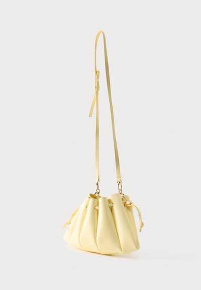 Απαλή κίτρινη δερμάτινη τσάντα τύπου κάδος με κορδόνι σύσφιξης, ρυθμιζόμενο λουράκι ώμου και χρυσούς μεταλλικούς εξαρτήματα σε απλό φόντο.