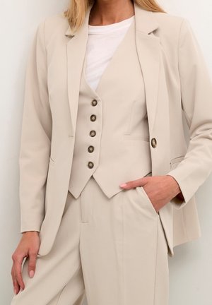Frau trägt einen beigen Anzug mit geknöpfter Weste, Blazer und Hose, Hand in der Tasche, darunter ein weißes Top.