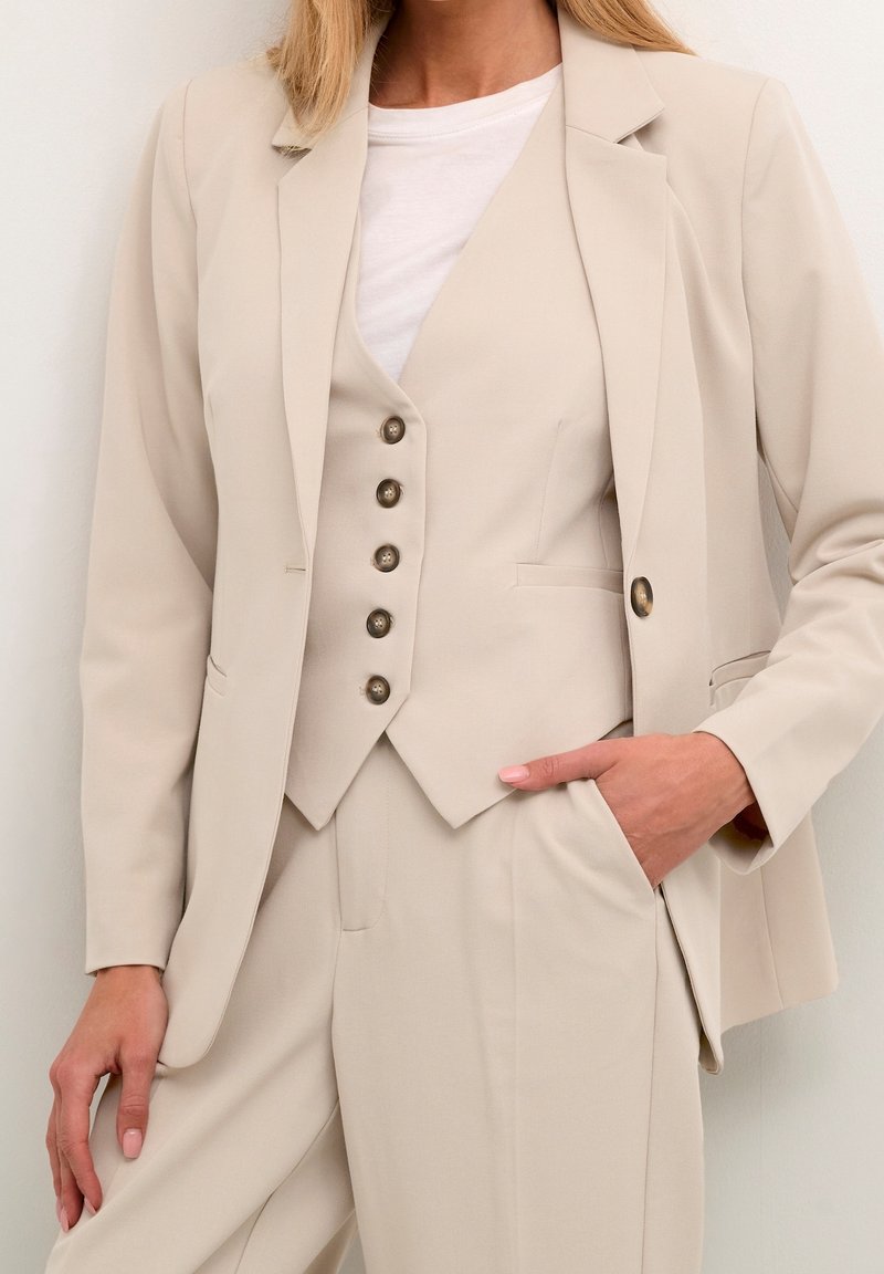 Femme portant un costume beige avec gilet boutonné, blazer et pantalon, main dans la poche, top blanc en dessous.