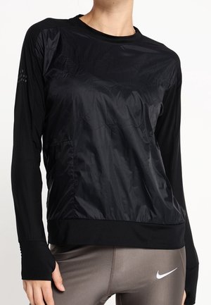 Frau trägt ein schwarzes, langärmliges Sportoberteil mit Daumenlöchern und graue Nike-Leggings, steht vor einem weißen Hintergrund.