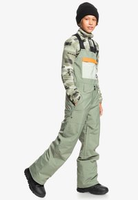 Groene sneeuwbroeken met verstelbare banden, functionele zakken en een oranje borstzak, gecombineerd met een gestippeld fleece shirt en een zwarte beanie.