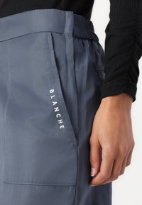 Graue, leichte Hose mit elastischem Bund, Seiten Tasche und dem Wort "BLANCHE" in weißer Stickerei im Bereich der vorderen Tasche.