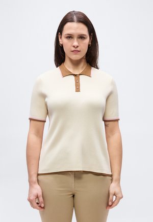 Ženska z ravnimi rjavimi lasmi, oblečena v kremasto polo majico s kratkimi rokavi in rjavim ovratnikom ter bež hlače, stoji pred enobarvnim svetlim ozadjem.