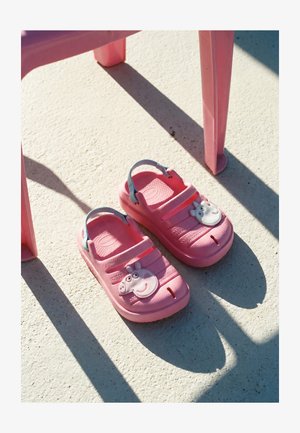 Paire de sabots roses pour enfants avec des visages d'animaux dessinés, placée sur un sol texturé sous une chaise rose projetant des ombres.