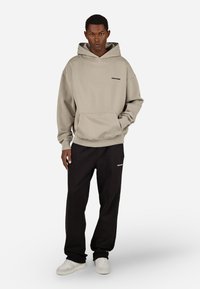 Beżowa bluza z kapturem z kieszenią z przodu, połączona z luźnymi czarnymi dresami z logo, uzupełniona białymi sneakersami.