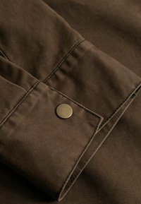 Κοντινό πλάνο καφέ μανσετώ ύφασμα με ορατή ραφή και ένα στρογγυλό ορειχάλκινο κουμπί με τριπλοκλίπ.