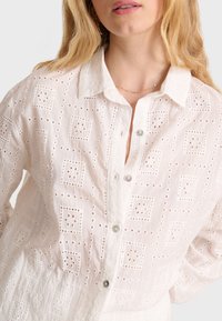 Camisa blanca de botones con patrones cuadrados bordados y detalles de ojal. Presenta un cuello y botones transparentes, confeccionada en una tela ligera.