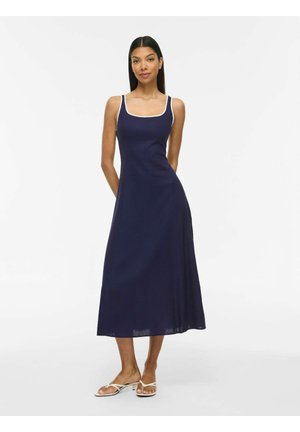 Femme portant une robe midi sans manches bleu marine avec bordure blanche et des sandales blanches, debout les mains dans le dos sur un fond blanc.