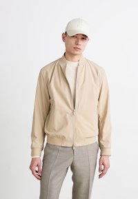 Beige bomberjakke med ribbestrikkede mansjetter og liv, med glidelås. Modellen har på seg en lys genser og en matchende caps.
