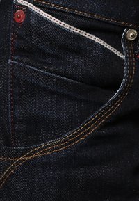 Los jeans de mezclilla oscura presentan un primer plano de un bolsillo delantero con costuras en blanco y naranja contrastantes, un botón de hardware rojo y sutiles detalles de textura.